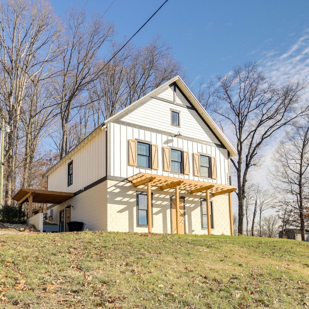 Modern farmhouse with white siding and wooden pergola on sloped green lawn surrounded by bare trees.